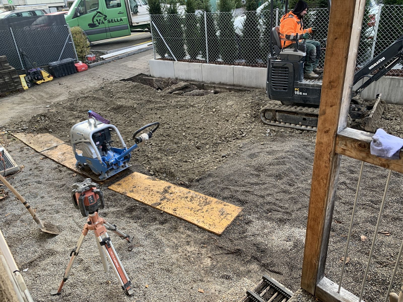 Baustelle mit Kleinbagger und Rüttelplatte vor einem Holzzaun